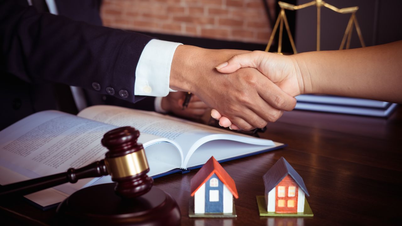 A lawyer and client shake hands, symbolizing a successful real estate deal finalized, with legal documents, a gavel, and miniature houses present on a dark wooden desk.