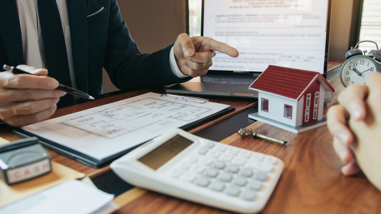 The image shows a close-up view of a wooden desk where a real estate agent and a client are seated. The agent, dressed in a dark suit, is pointing at a house blueprint spread on the desk. The blueprint depicts the floor plan of a house.
